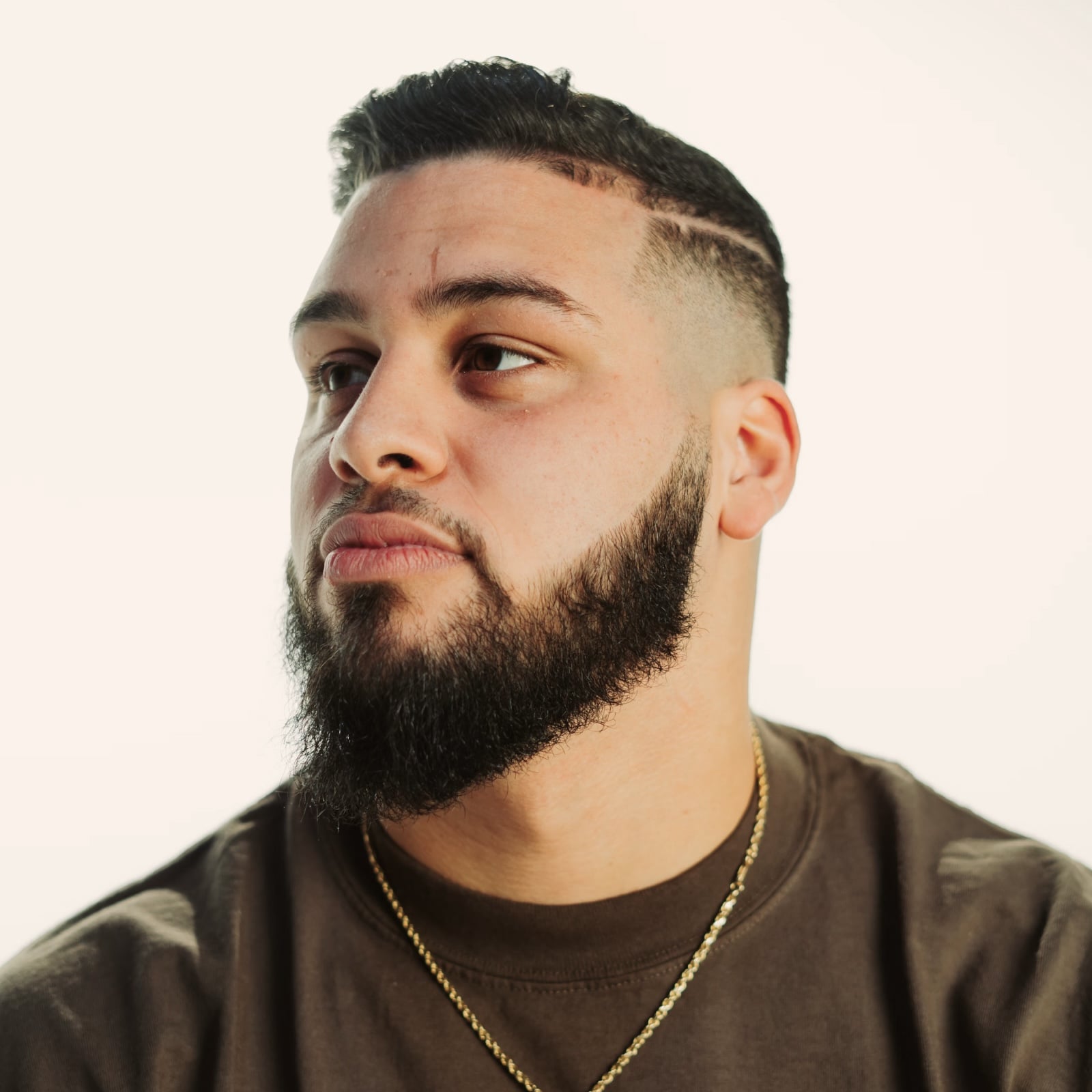 Person with styled hair and beard looking sideways, wearing gold necklace